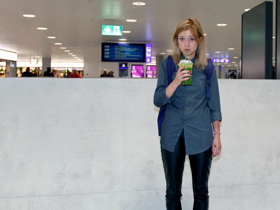 Eine junge Frau mit betrübtem Blick in einer Bahnhofshalle. Sie trinkt einen grünen Saft aus einem Becher.