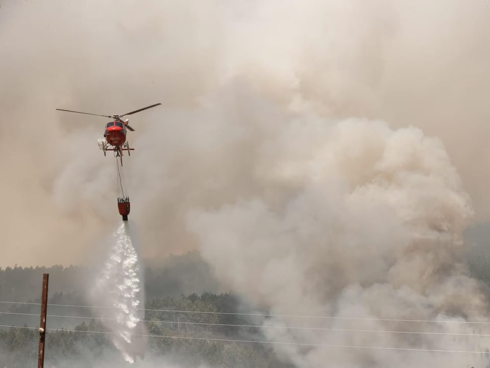 Ein Löschhelikopter fliegt vor einer Wand aus Rauch. Er wirft Wasser ab.