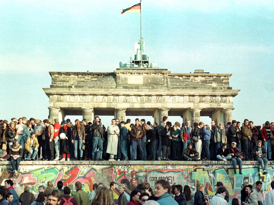 Viele Menschen stehen vor und auf der Berliner Mauer. Im Hintergrund das Brandenburger Tor.