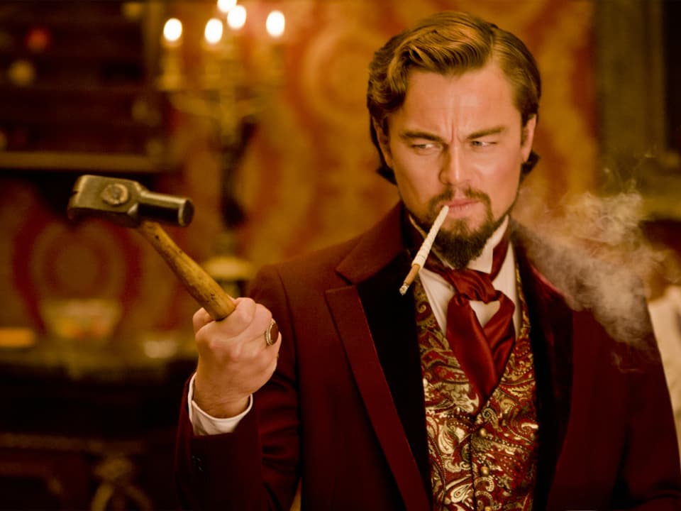 Leonardo DiCaprio hält in schicker Kleidung und mit einer Zigarette im Mund einen Hammer in der Hand.