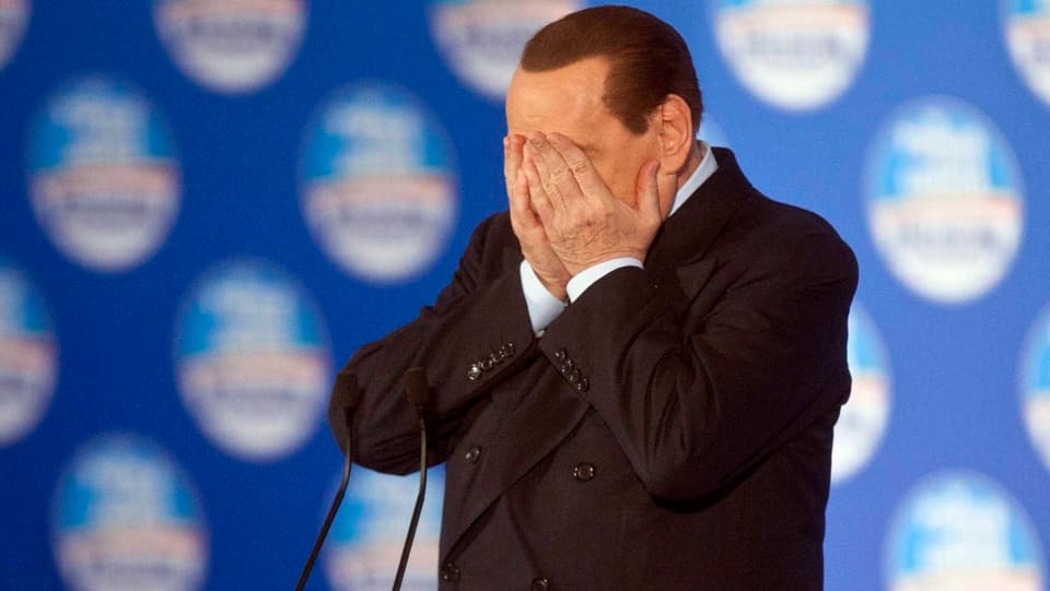 Silvio Berlusconi steht hinter einem Rednerpult und schlägt beide Hände vor das Gesicht.