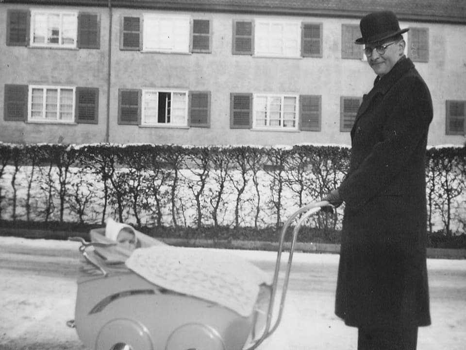Ein Mann mit Hut und langem Mantel vor stösst einen Kinderwagen, in dem ein Baby mit einer Mütze sitzt.