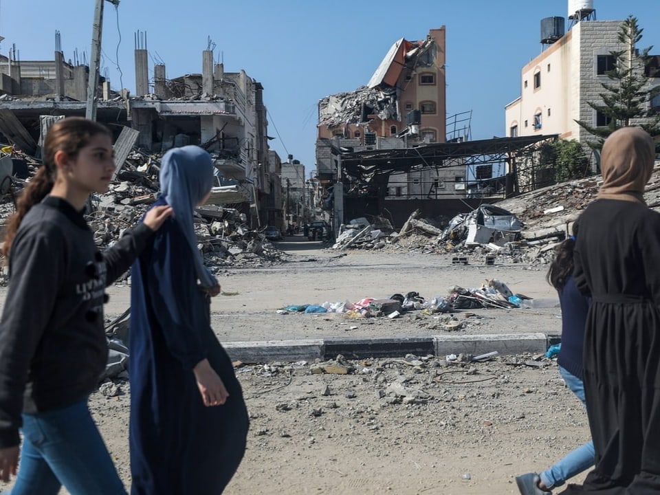 drei Frauen laufen an Ruinen von Gebäuden entlang, zwei davon mit Kopftuch