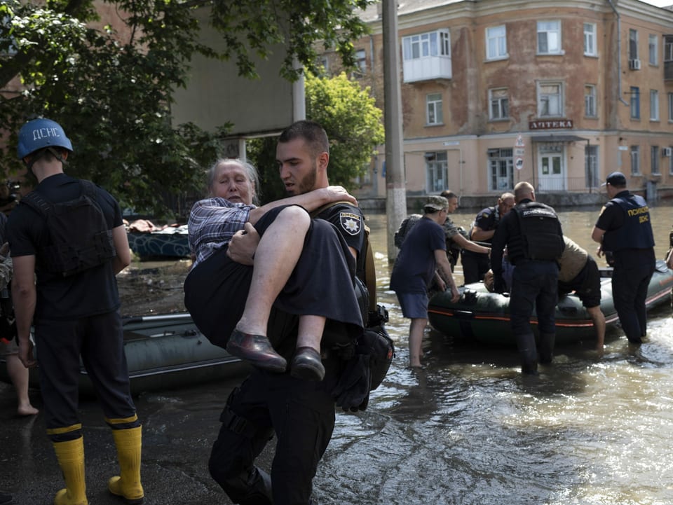 Ein Mann trägt eine alte Frau von einer überschwemmten Strasse weg. Im Hintergrund ist ein Schlauchboot mit Helfern.