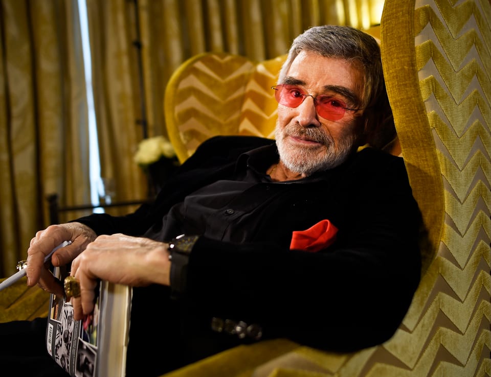 Burt Reynolds liegt auf einem Sofa und trägt eine Sonnenbrille mit roten Gläsern.