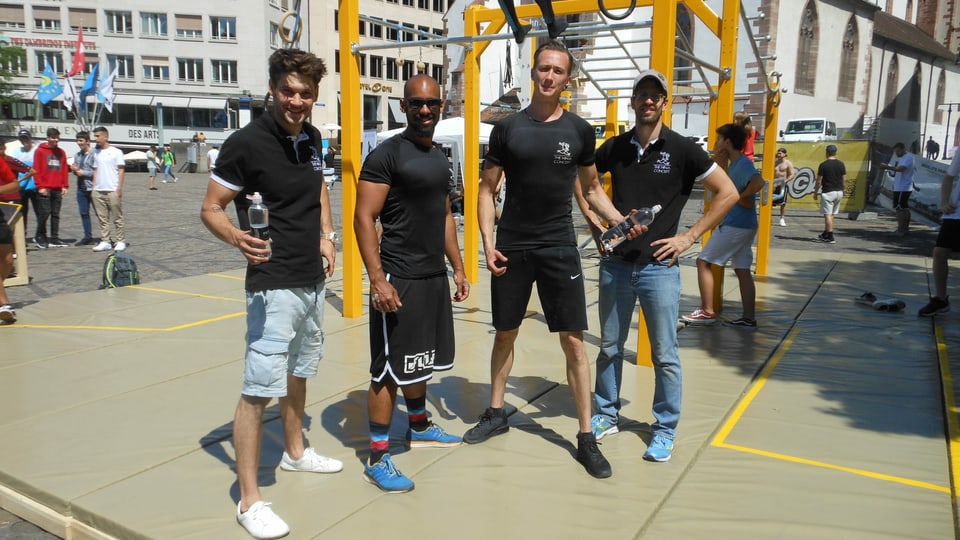 Vier Sportler in schwarzen T-Shirts posieren vor einem gelben Klettergerüst.