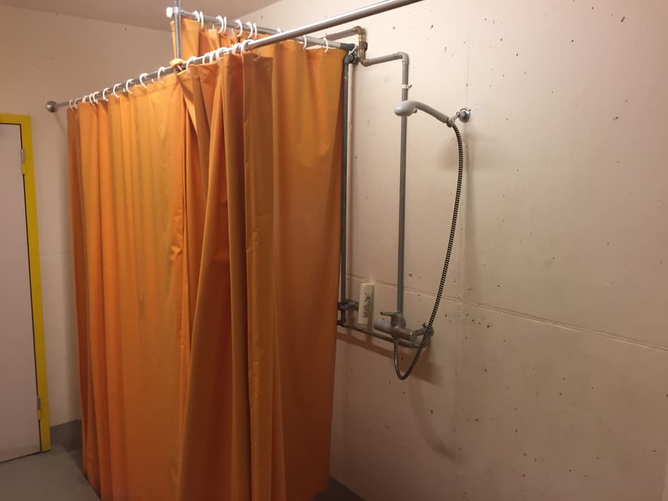 Duschvorhänge (Orange) vor einer weissen Wand mit Leitungen. 