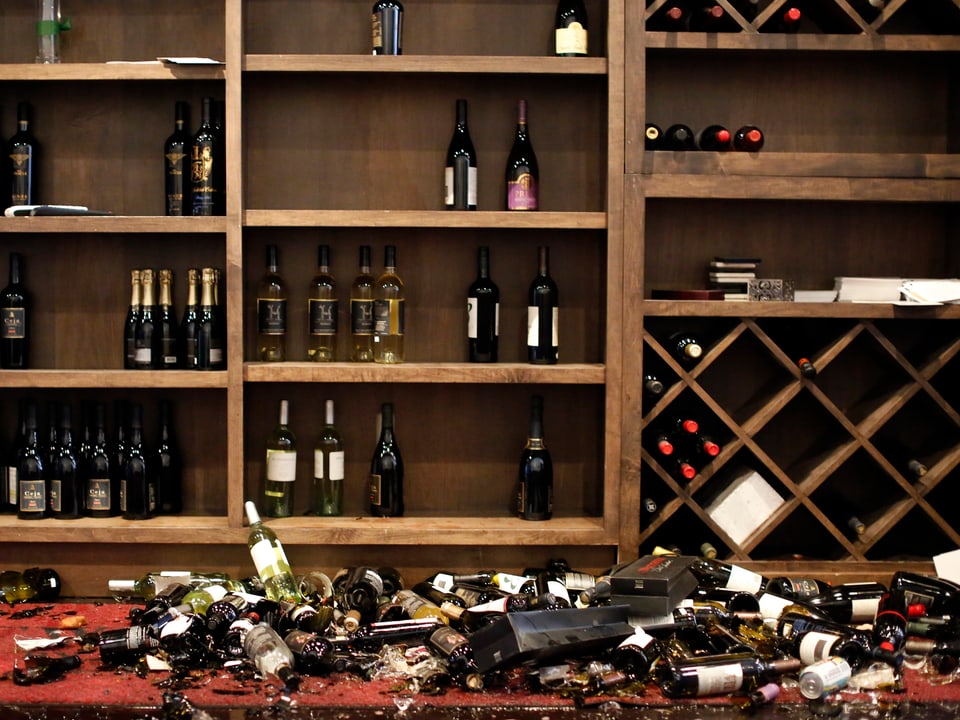 zerborstene Weinflaschen liegen vor einem Weingestell am Boden