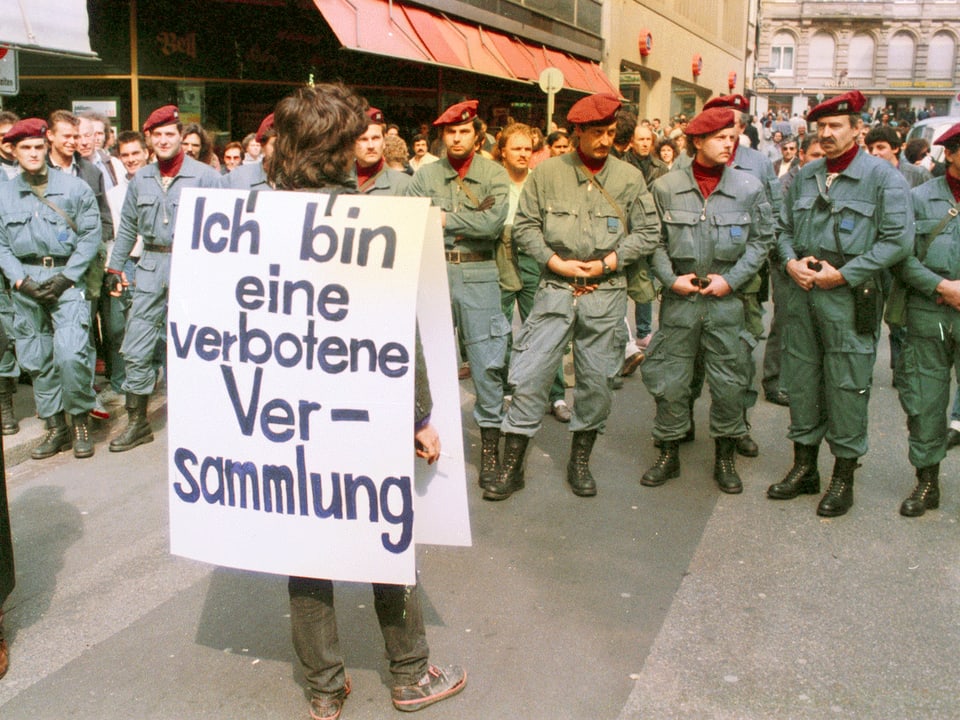 1989: Ein Mann mit einem Schild mit der Aufschrift «Ich bin eine verbotene Versammlung» steht vor einer Reihe Polizisten.