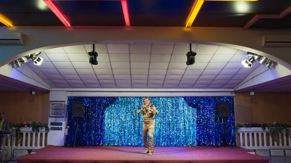 Foto einer Bühne mit rotem Teppich, blau, glitzernder Wand. Darauf steht ein Mann in goldenem Anzug und singt.
