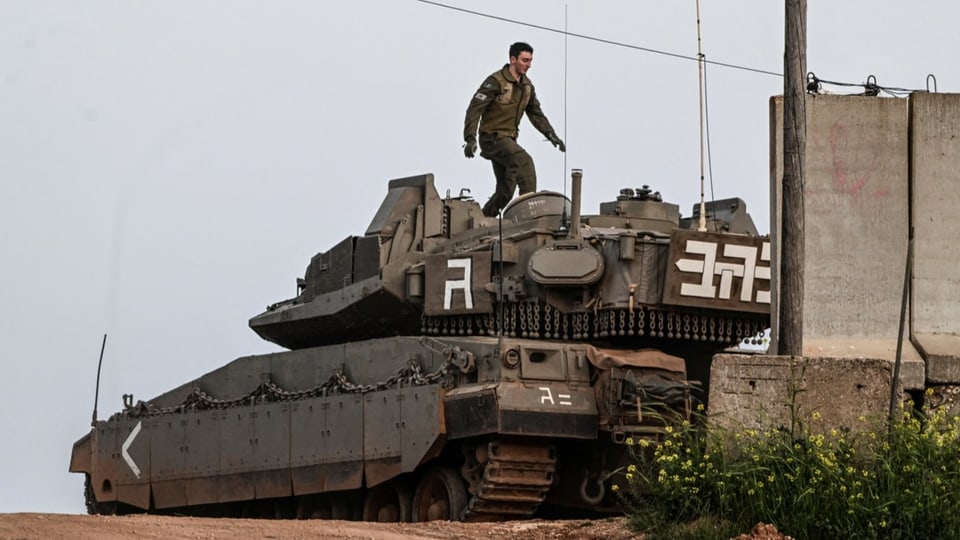 Ein Soldat steht auf einem Panzer. Im Bild ist eine Mauer und eine Stromleitung zu sehen.