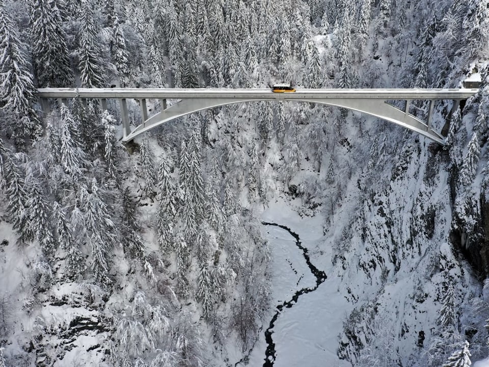 Verschneites Tal mit Wald und Betonbrücke. Auf der Brücke ein gelbes Postauto.
