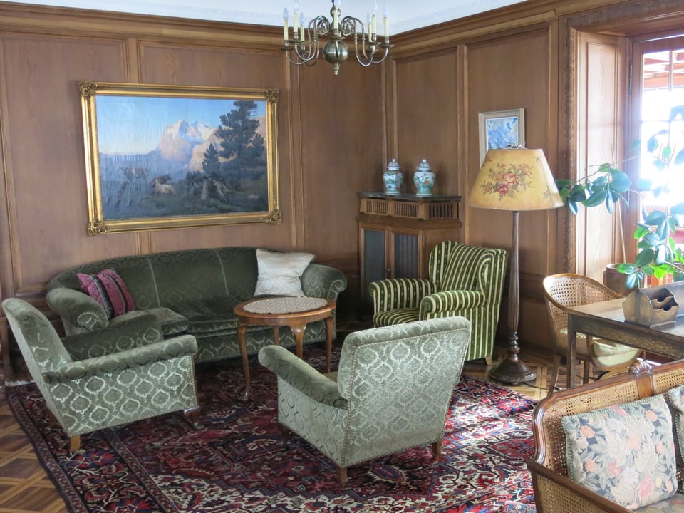 Der Salon mit grünen Polstermöbeln, grossem antikem Bild und Kronleuchter.