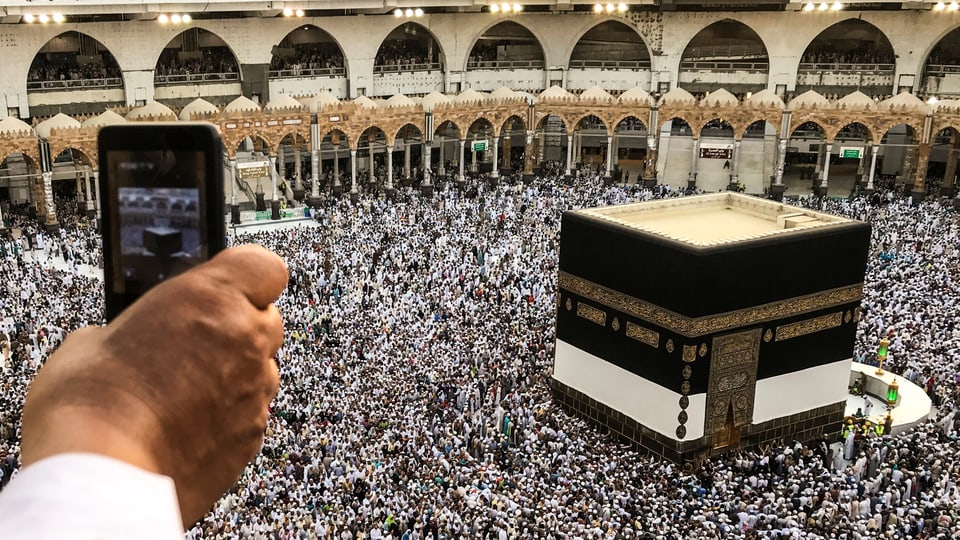 Eine Hand hält ein Smartphone, im Hintergrund ist die Kaaba in Mekka zu sehen.
