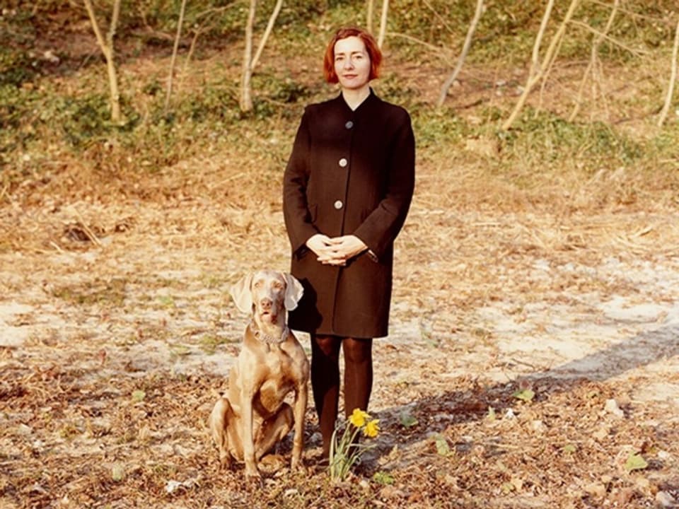Eine Frau mit kurzen, roten Haaren und einem schwarzen Mantel steht am Waldrand, neben ihr ein Hund.