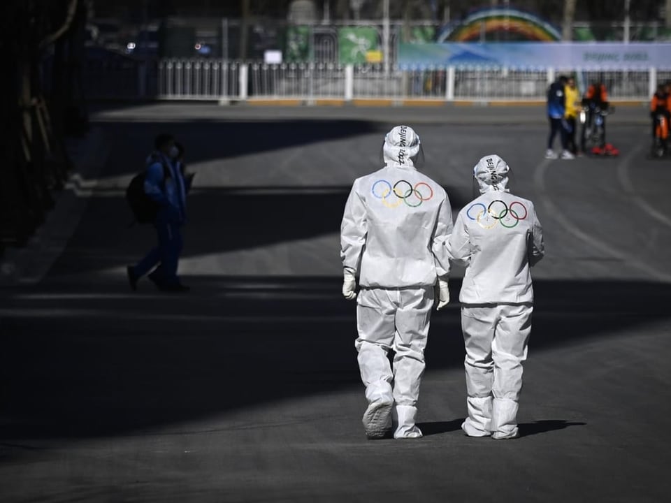 Zwei Personen in Schutzanzug mit den Olympischen Ringen auf dem Rücken gehen vorbei.