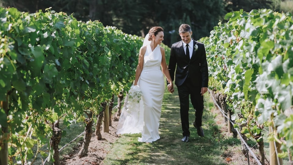 Das Paar ist Hand in Hand zwischen Reihen von Weinstöcken zu sehen.