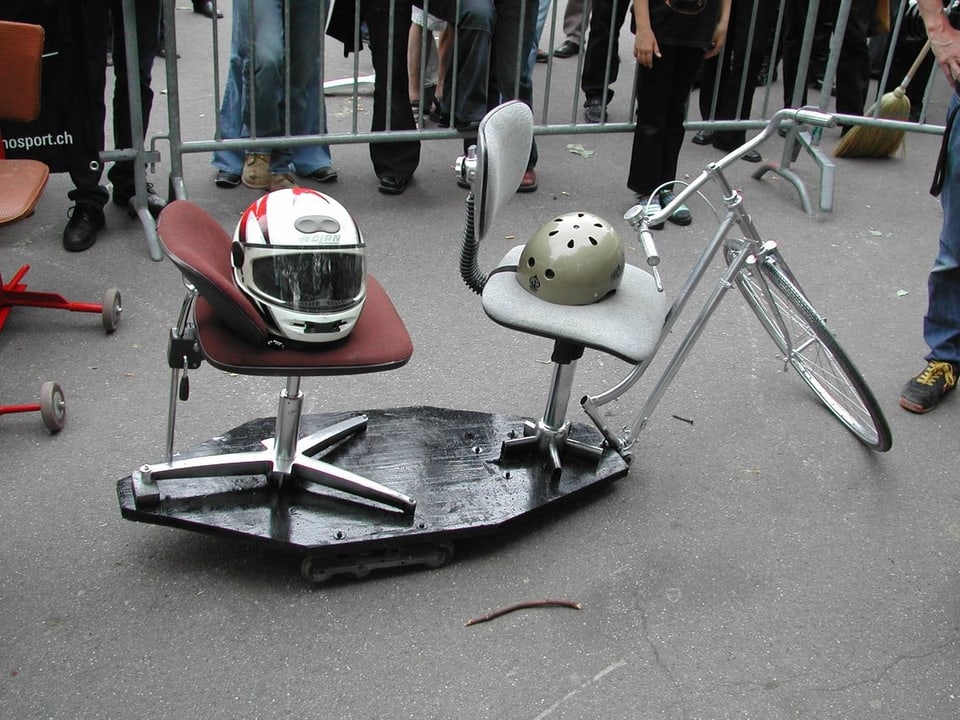 Zwei Bürostühle, fest gemacht auf einer Holzplatte mit rädern, davor die vodere Hälfte eines Velos mit Lenker und einem Rad.