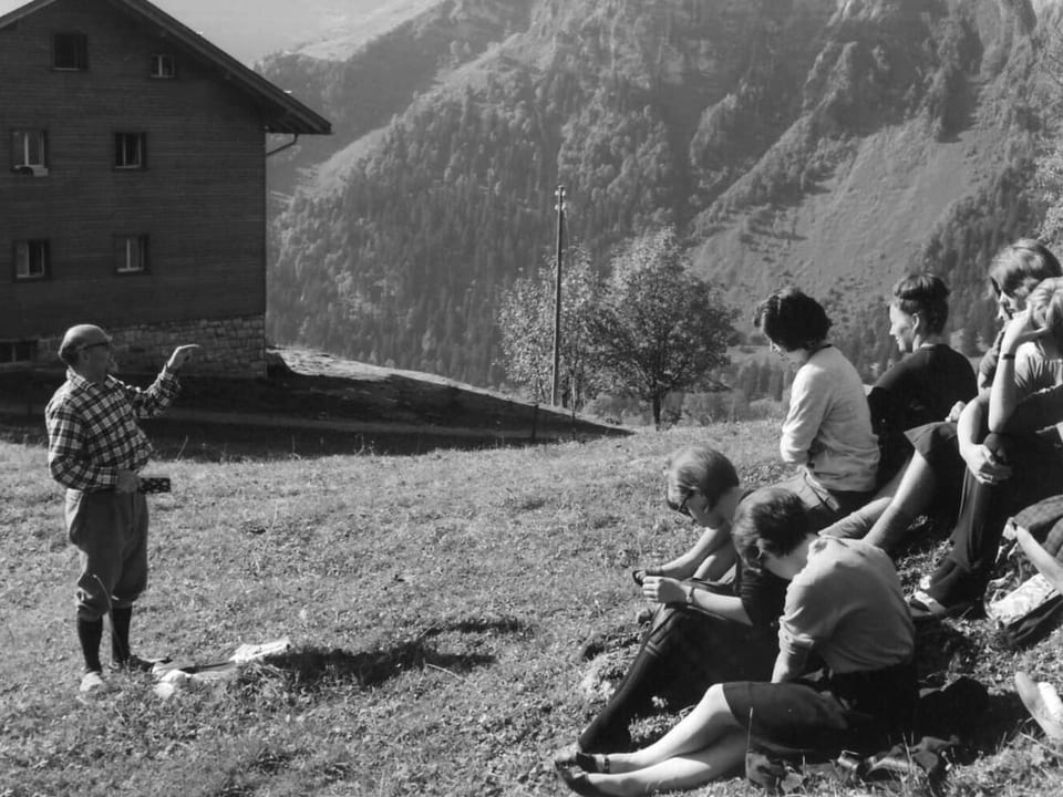 Jugendliche sitzen auf einem Hügel, ein Erwachsener spricht zu ihnen. 