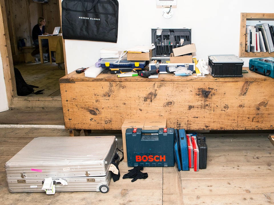 Werkzeuge und Koffer liegen am Boden und auf einer Kommode, im Hintergrund ein Mann am Laptop