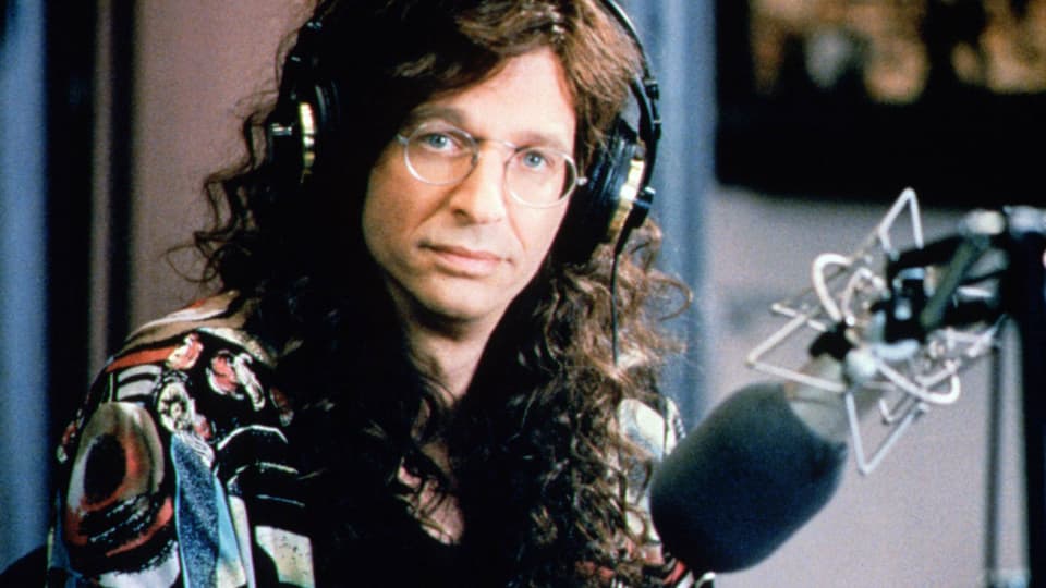 Ein Mann mit Brille und langen Haaren sitzt vor einem Mikrophon und schaut in die Kamera.