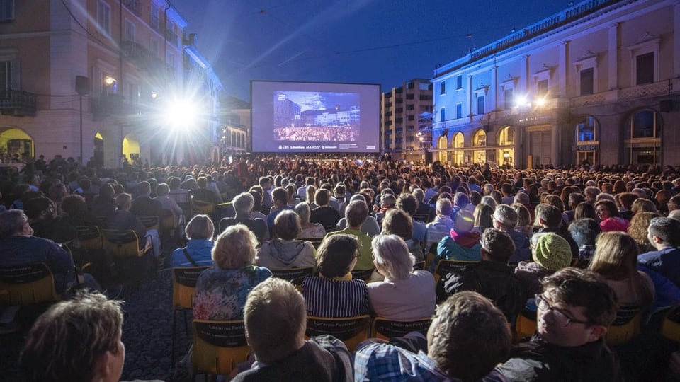 Ein Open-Air-Kino auf einem Platz mitten in der Stadt, alle Stühle vor der Leinwand sind besetzt.