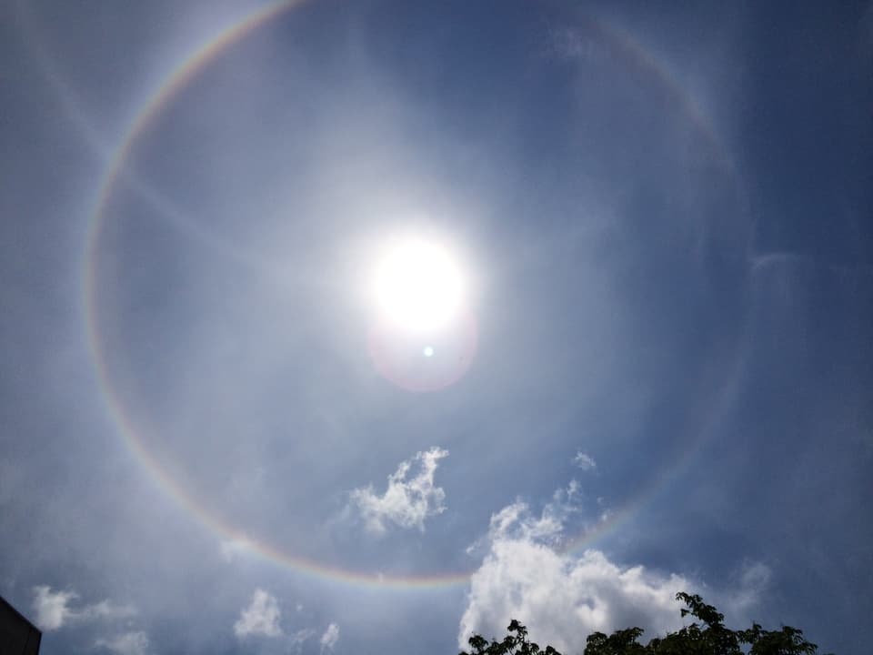 Milchig-blauer Himmel mit Sonnenscheibe in der Mitte. Um die Sonne ist ein feiner Kreis in allen Regenbogenfarben zu sehen. 