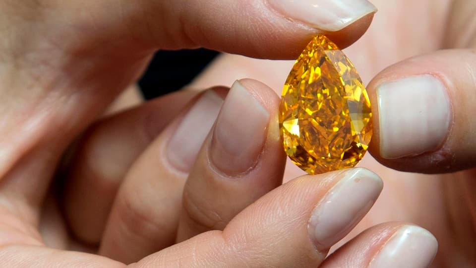Der orange farbene Diamant zwischen Damenfingern. Seine Form jene eines Tropfens. 