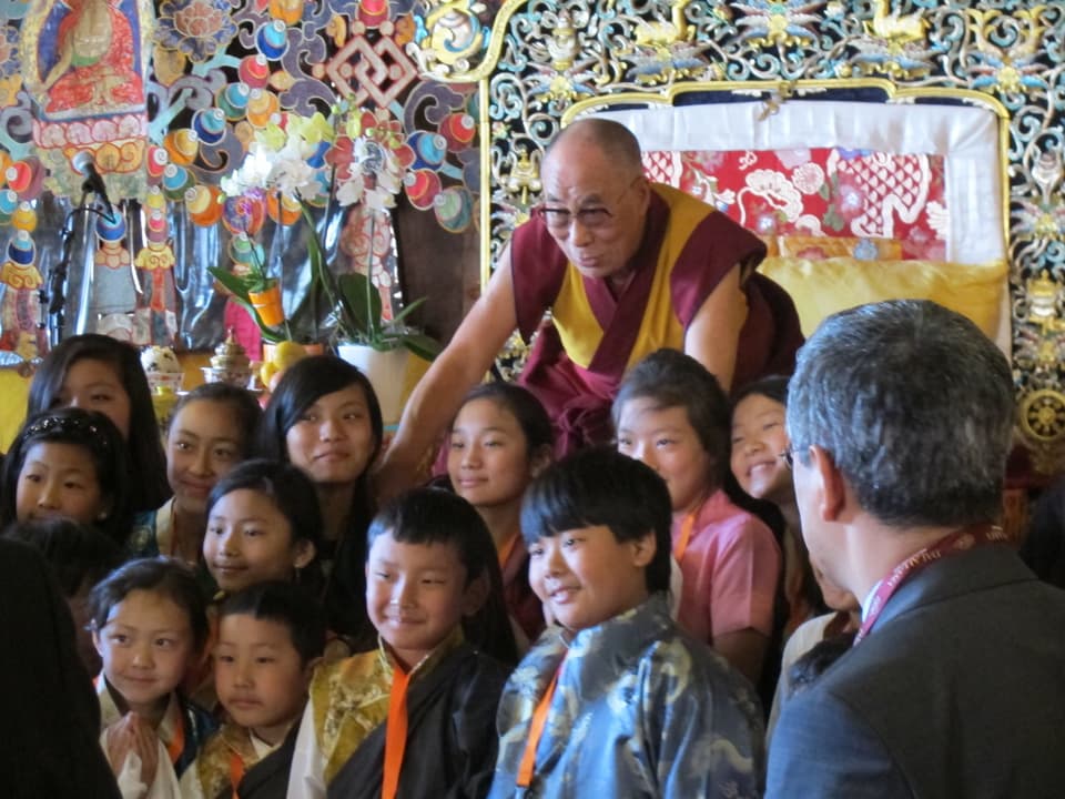Der Dalai Lama beugt sich zu tibetischen Kindern und Jugendlichen hinunter.
