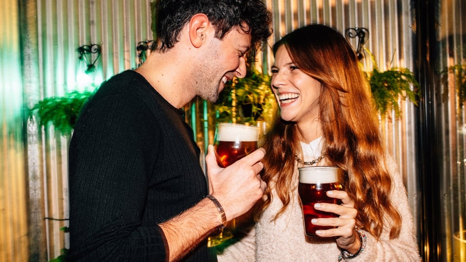 Ein Mann und eine Frau schauen sich verliebt in die Augen und lachen, sie halten beide ein Bier in der Hand.