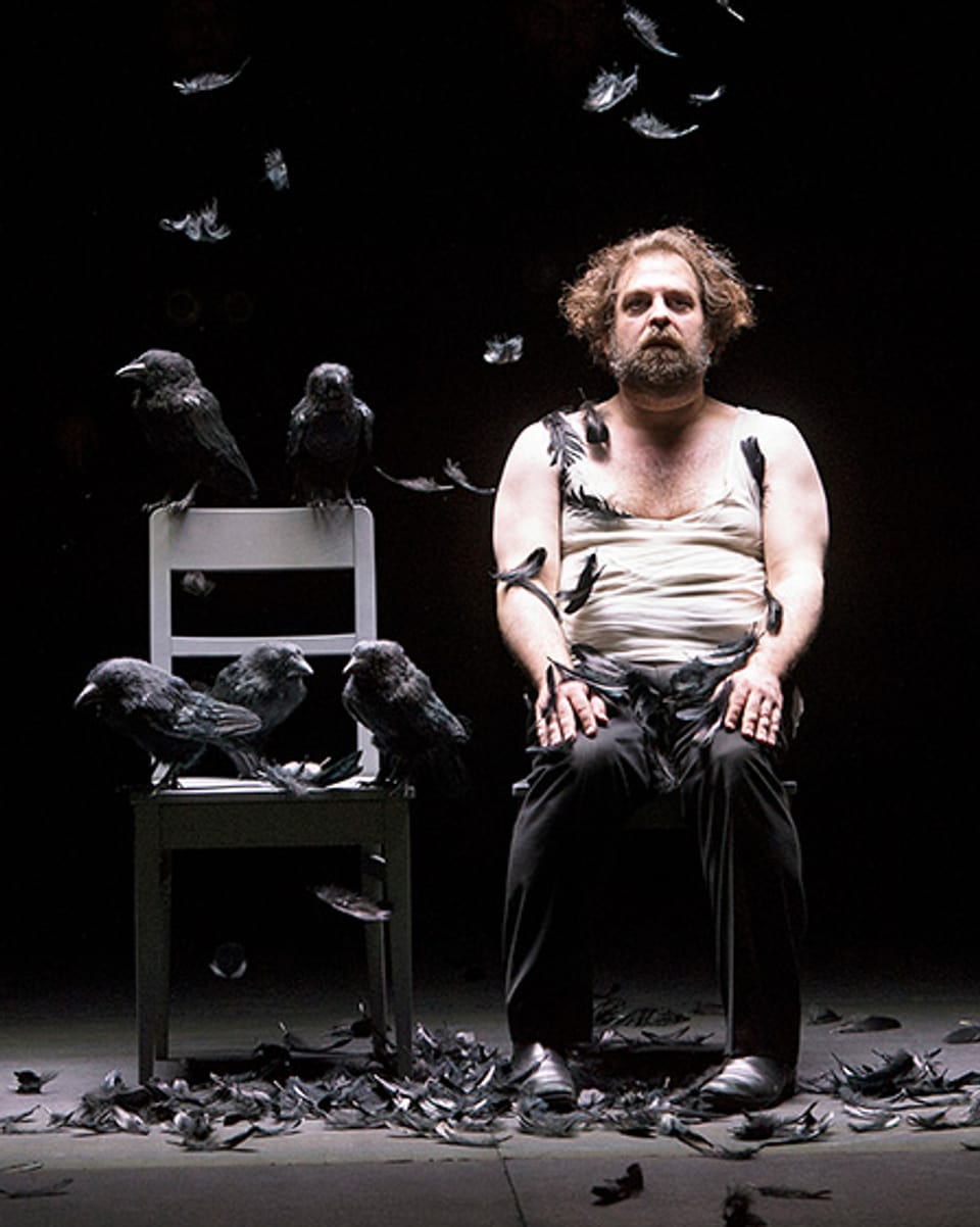 Ein Mann sitzt auf einer Bühne. Auf dem Stuhl neben ihm hocken schwarze Vögel.