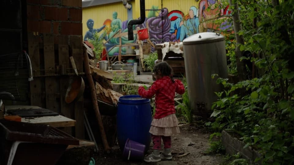 Kleines Kind steht neben einer Wassertonne, im Hintergrund ein bemalter Bauwagen