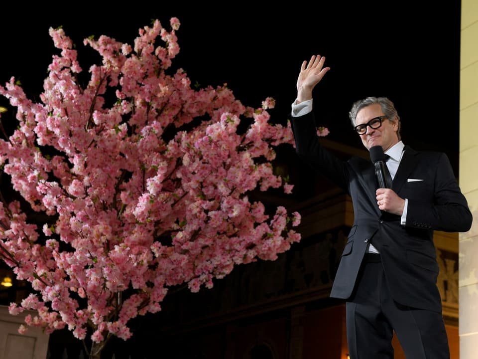 Colin Firth steht vor einem Baum mit rosaroten Blüten und hält ein Mikrofon in der linken Hand.