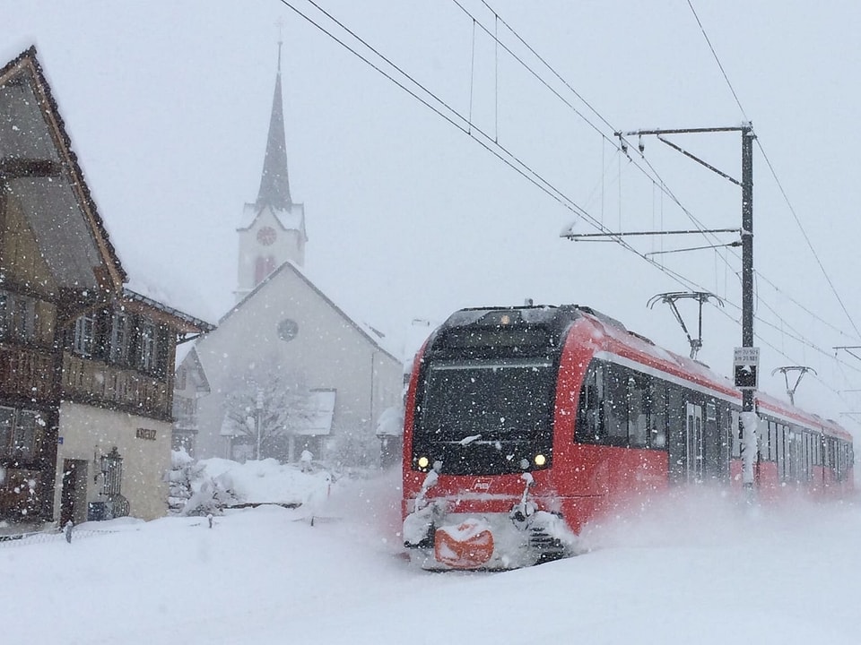 Eine Rote Lokomotive der Appenzellerbahn fährt durch das Schneegestöber. Im Hintergrund steht eine Kirche.