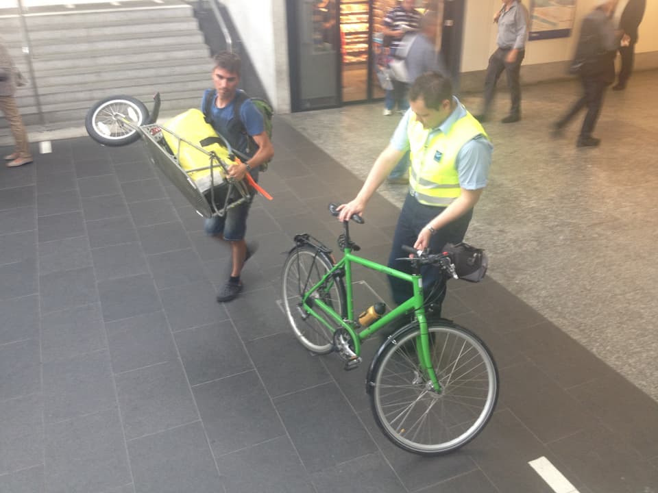 Securitrans-Mitarbeiter nimmt ein grünes Herren-Fahrrand. Der Besitzer daneben trägt den Veloanhänger mit dem Gepäck. 