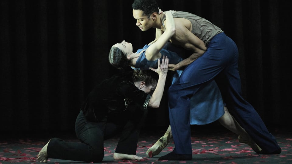 Drei Tänzer führen eine zeitgenössische Tanzszene auf dunkler Bühne auf.