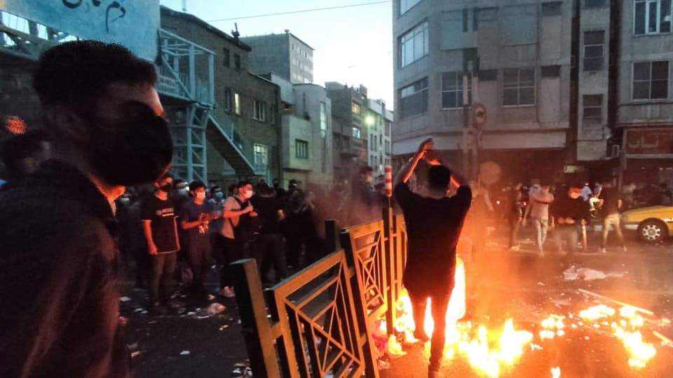 Menschen auf den Strassen Irans. Flammen und Barrikaden. Sie tragen Gesichtsmasken. Es ist halbdunkel.