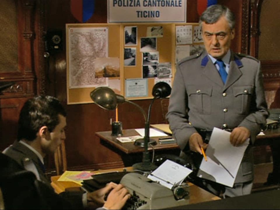 Polizist in grauer Uniform in einem Büro. Im Hintergrund ein Schild mit der Aufschrift "Polizia Cantonale Ticino"