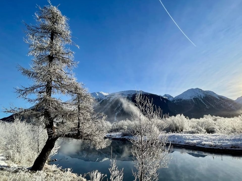 Samedan/GR Blick über Fluss auf verschneite Landschaft mit Reif. Sonne steht tief am blauen Himmel.