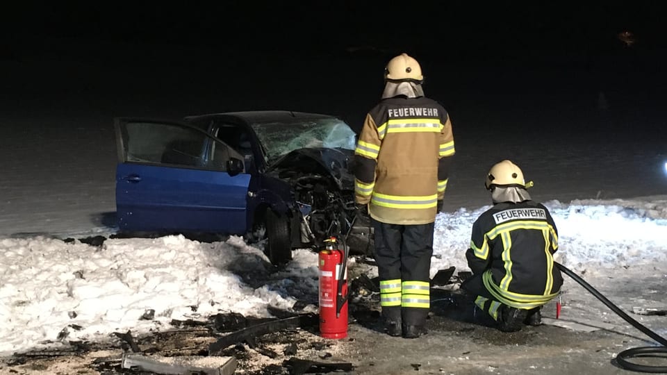 Zwei Feuerwehrmänner stehen, bzw. knien vor dem ausgebrannten Auto