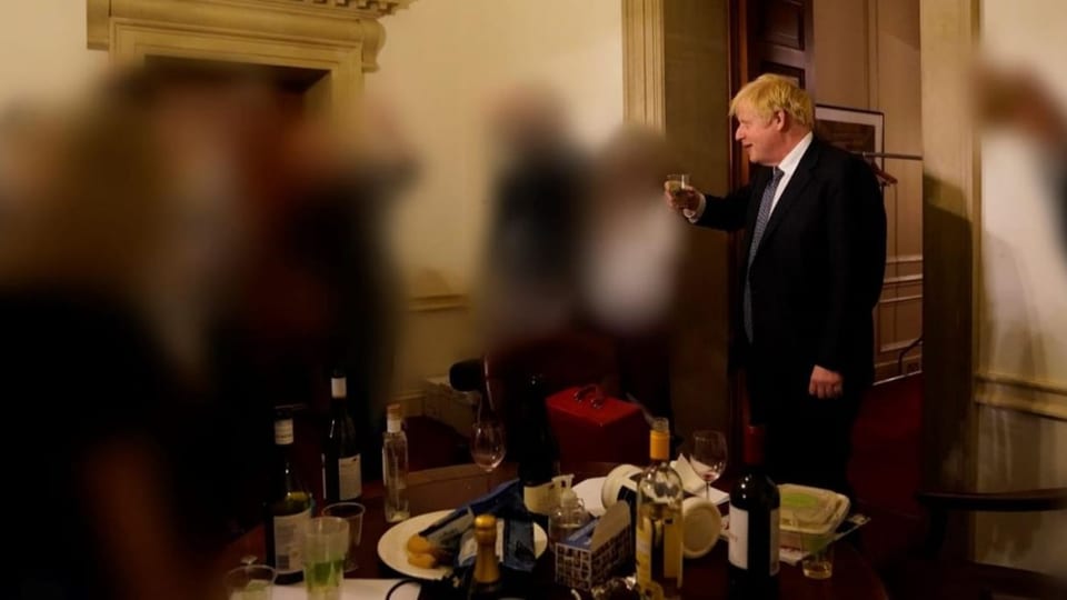 Boris Johnson mit einem Glas in der Hand, auf dem Tisch Flaschen und Snacks. Weitere Personen sind unkenntlich gemacht.