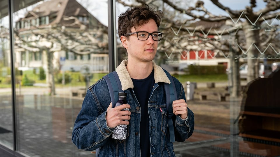 Ein Jugendlicher steht mit einem Rucksack vor der Glasfassade eines Gebäudes.