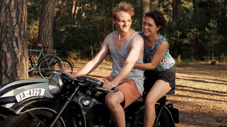 Ein junger Mann und eine junge Frau sitzen im Sommer auf einem Motorrad und lächeln.