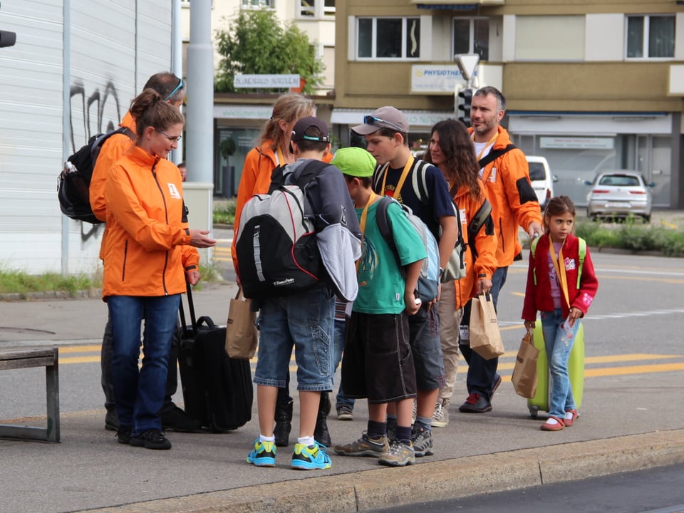 Familie Ambrosini und Familie Leonardi warten mit Gepäck und in orangen Jacken an der Tramhaltestelle.