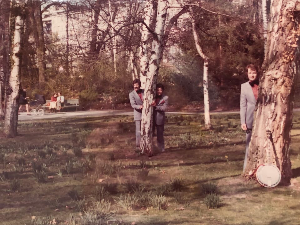 Drei Männer stehen hinter Bäumen in einem Park.