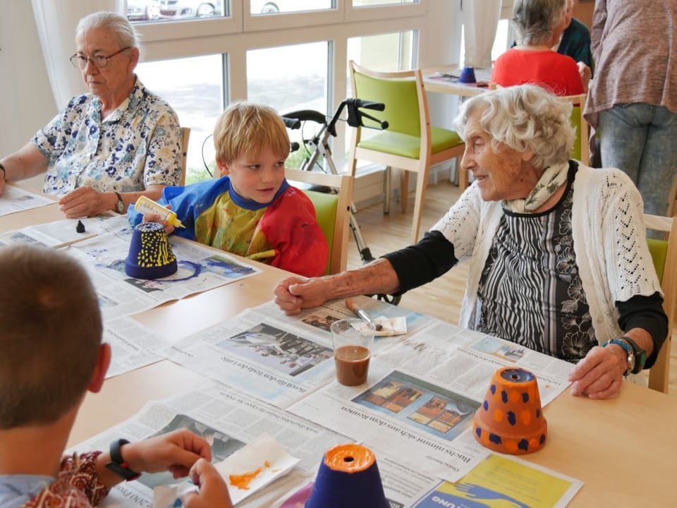Ein Bub und zwei Seniorinnen sitzen am Tisch und bemalen Tontöpfe