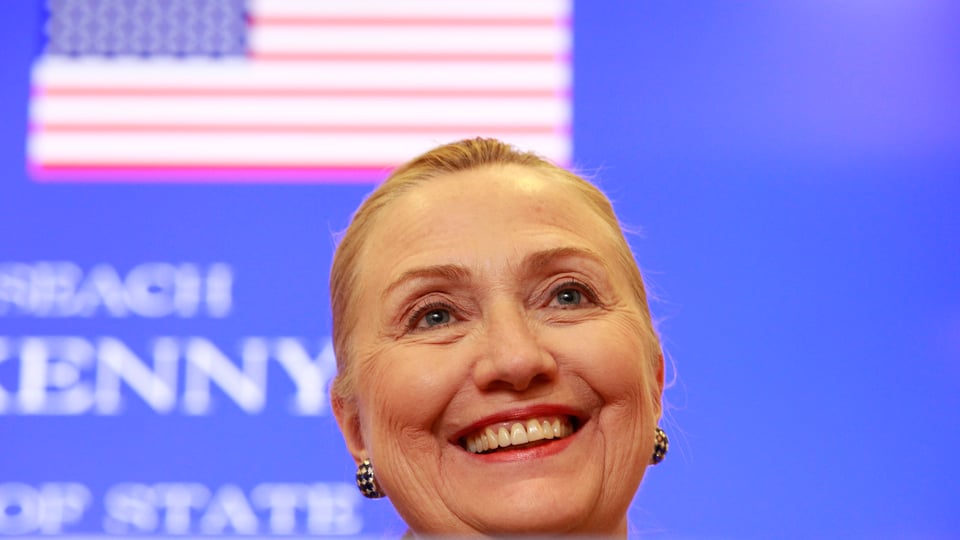 Clinton lächelnd vor blauer Wand mit US-Flagge.
