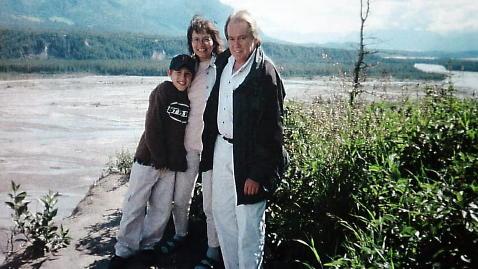 Vater, Mutter und Sohn haben sich die Arme um die Schulter gelegt. Sie stehen an einem Flussufer.