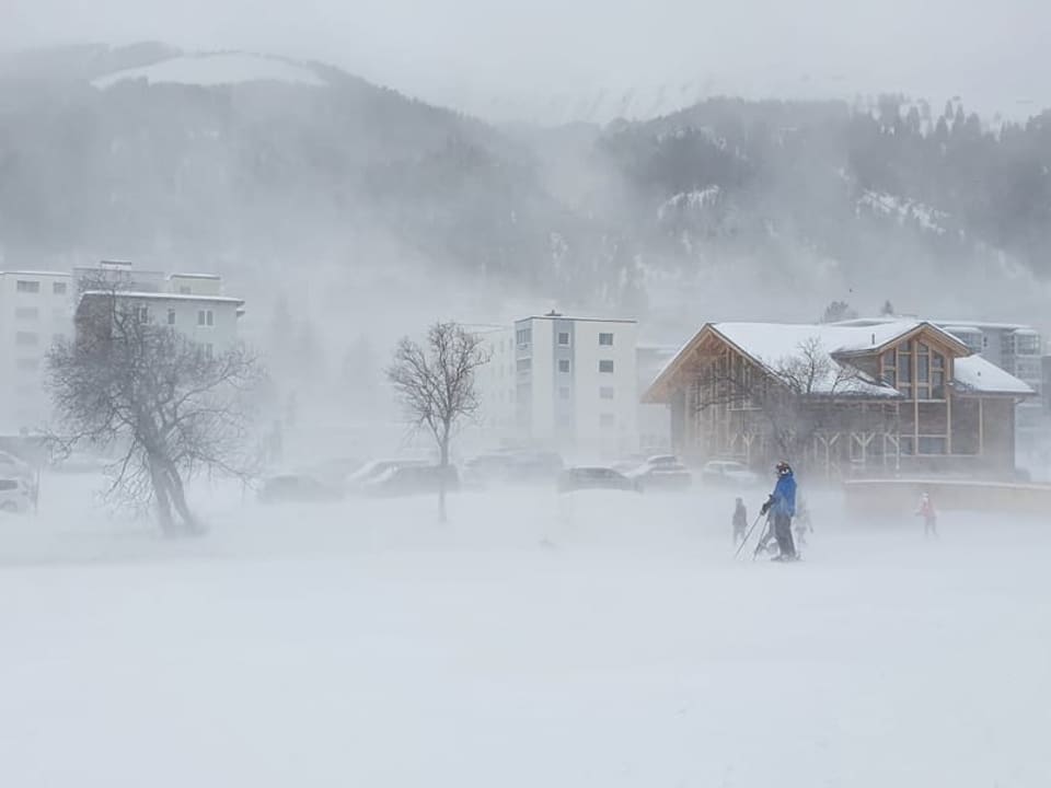 Schnee wird verweht, dazwischen stehen Personen in Skiausrüstung