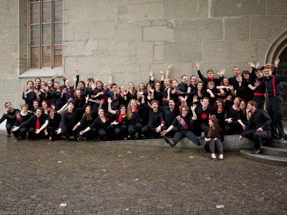 Die Sängerinnen und Sänger tragen schwarze Kleider mit roten Accessoires und posieren kniend für Gruppenfoto.
